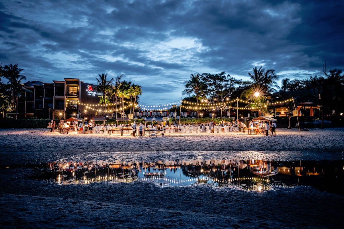 Beach celebration at dusk with string lights, tables, and guests gathered on the sand